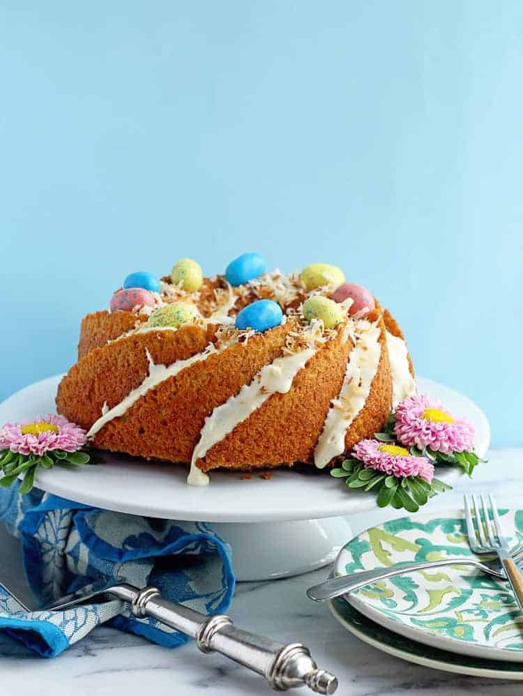 Carrot Cake Recipe in full display on a white cake plate topped with Easter egg shaped candies and coconut, surrounded by fresh flowers.