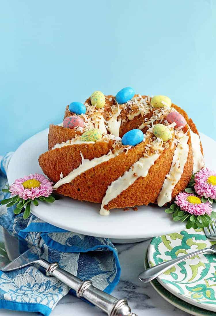 Easy Carrot Cake Recipe displayed on a white cake plate topped with Easter egg candies and coconut. 