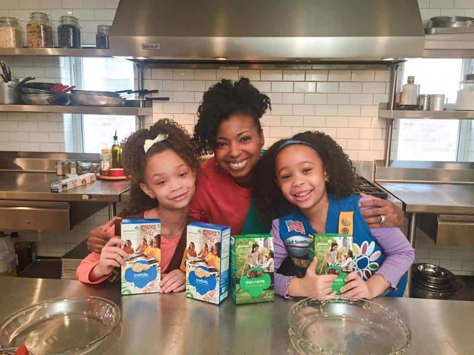 Jocelyn Delk Adams poses with two girl scouts and their boxes of cookies in a kitchen