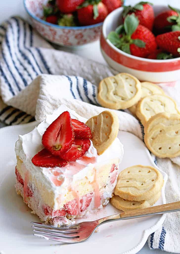 A piece of Girl Scout Cookies Strawberry Ice Cream Cake sitting on a white plate with trefoil cookies and topped with sliced strawberries and two bowls of fresh strawberries in the background