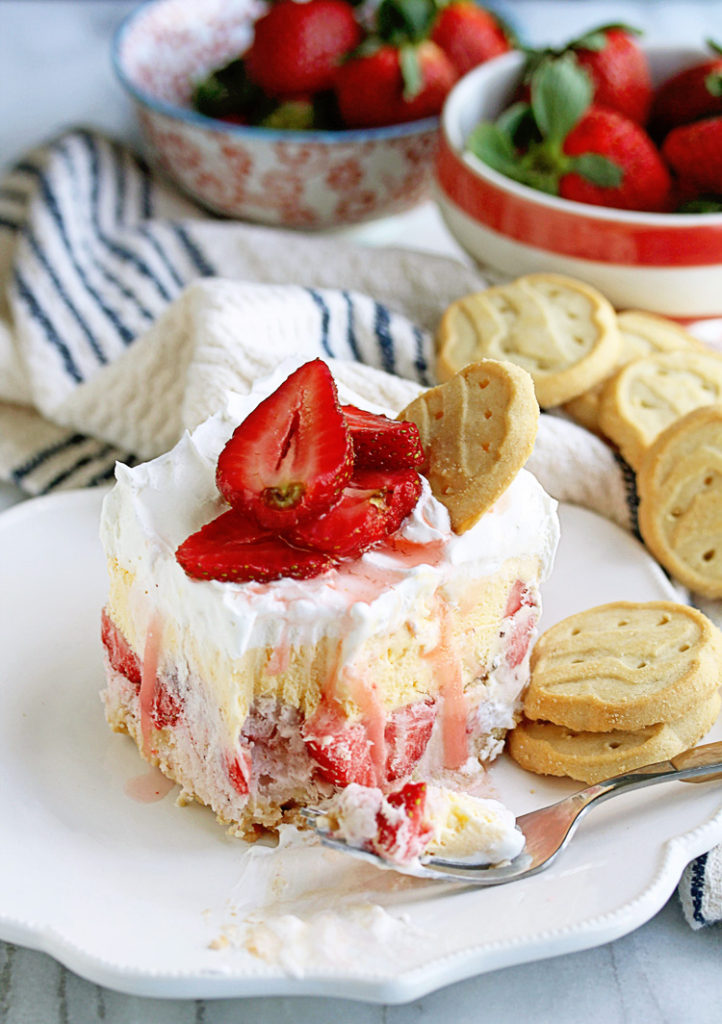 Close up of a piece of Girl Scout Cookies Strawberry Ice Cream Cake sitting on a white plate with trefoil cookies and topped with sliced strawberries and two bowls of fresh strawberries in the background