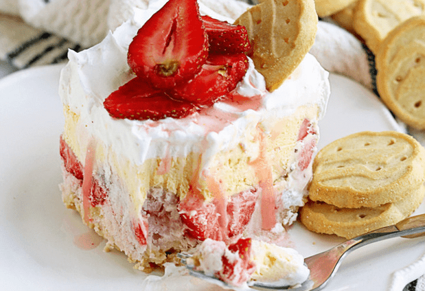 A piece of Girl Scout Cookies Strawberry Ice Cream Cake sitting on a white plate with a fork and topped with sliced strawberries and a trefoil cookie
