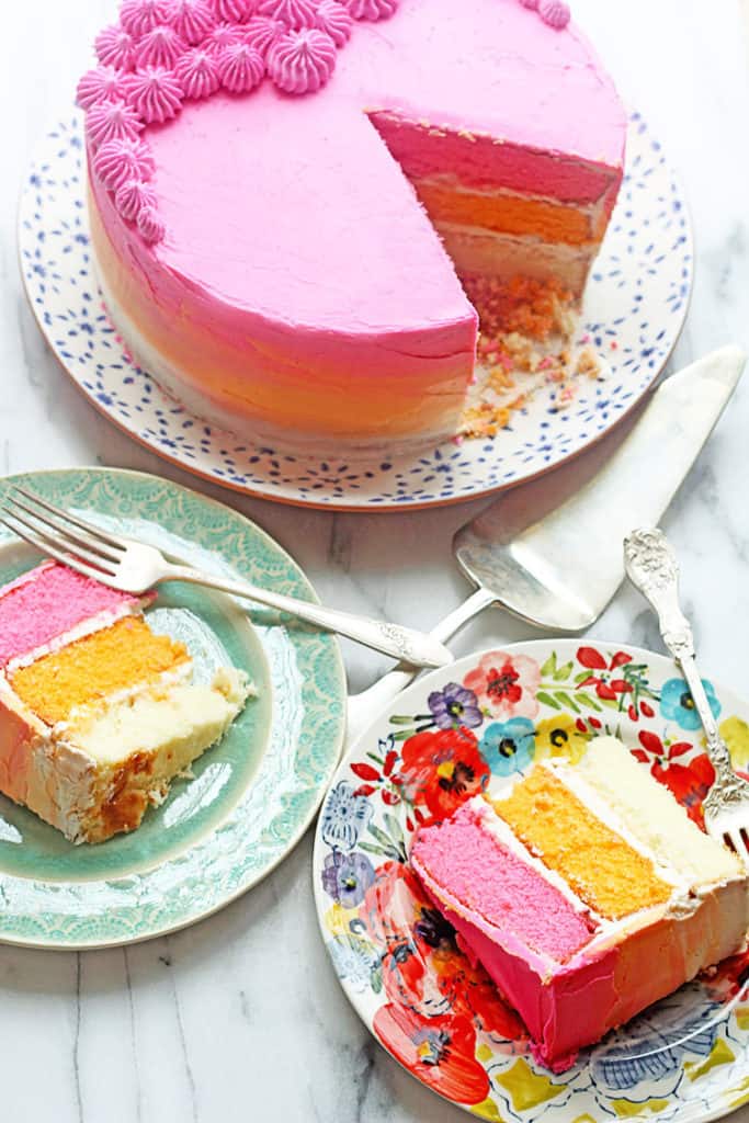 Two slices of a three layer ombre cake displayed on two different round plates with forks and the entire cake with slices missing next to them