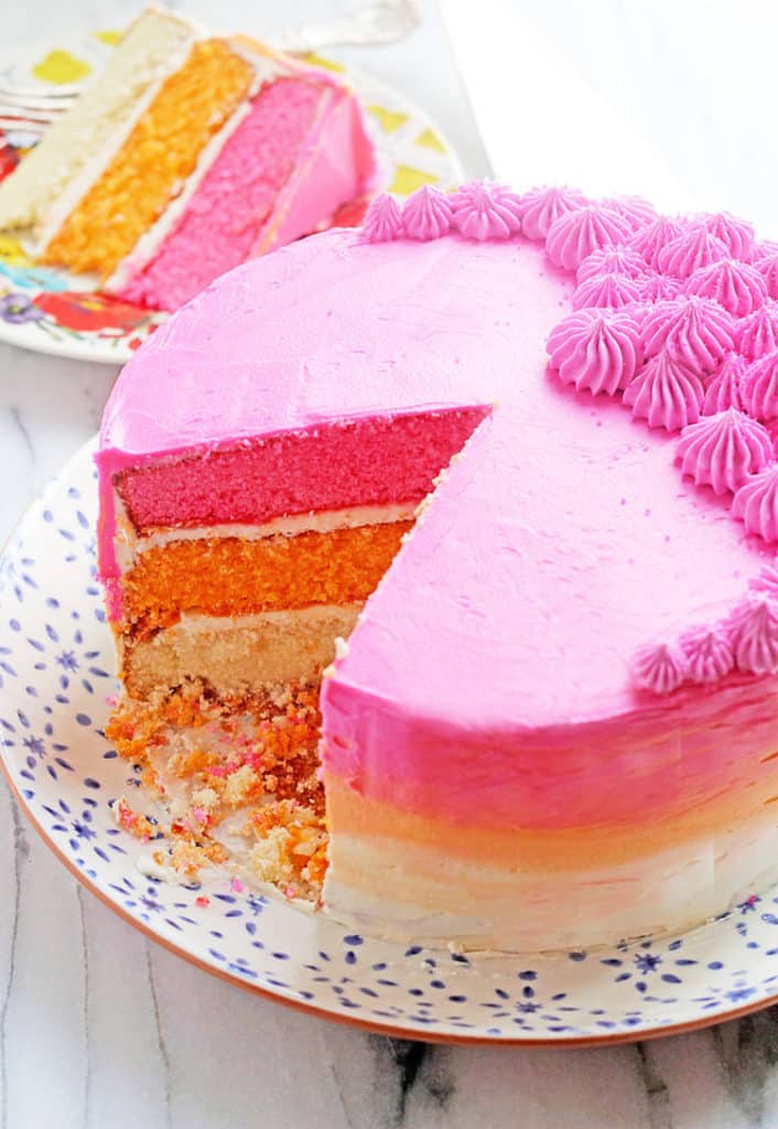 Beautiful three layer ombre cake with slices missing and a slice sitting on a colorful, round plate in the background