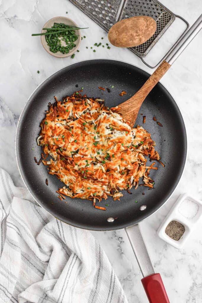 Cooked hash browns in a skillet on the counter with a potato and other ingredients around the pan.