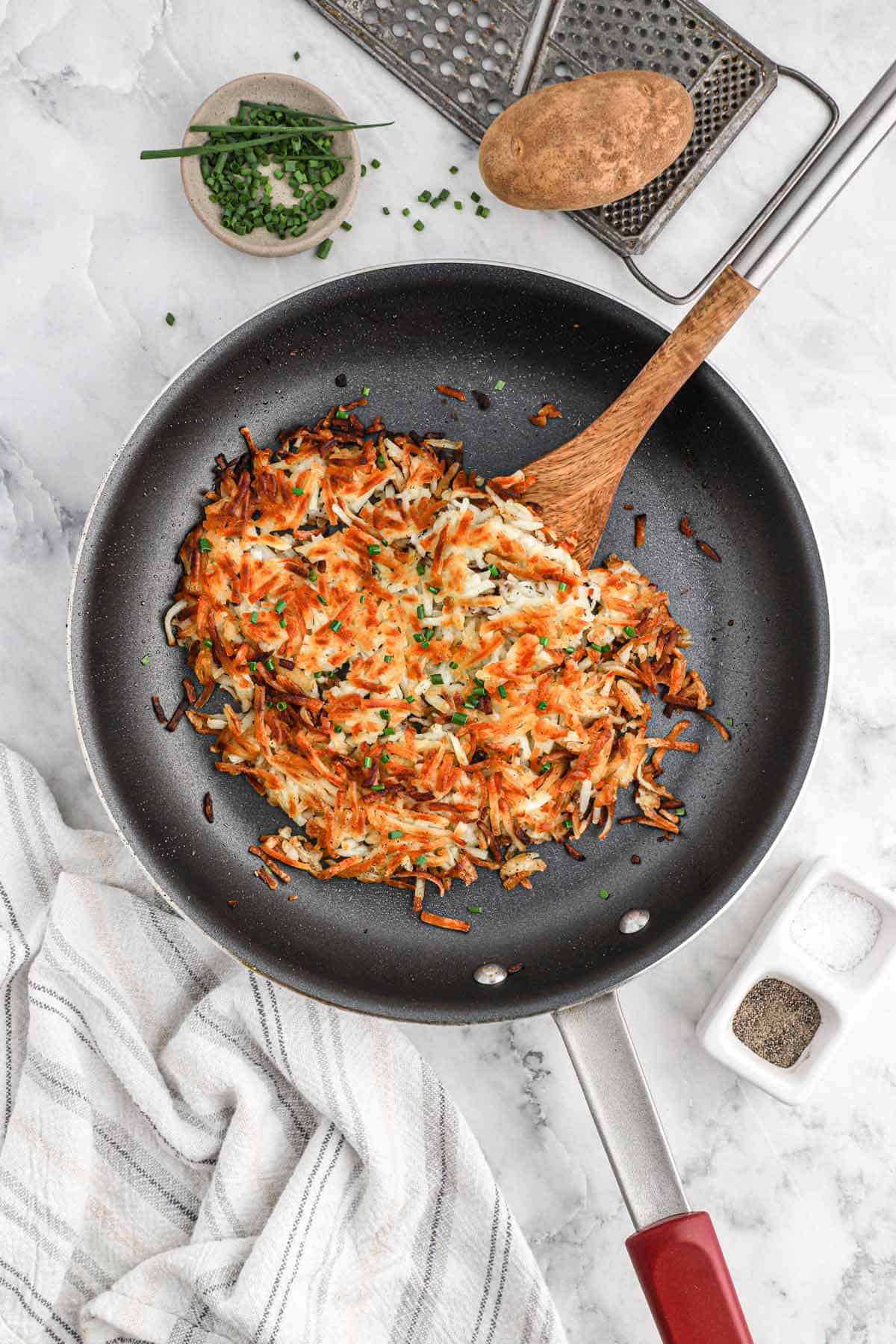 Cooked hash browns in a skillet on the counter with a potato and other ingredients around the pan.