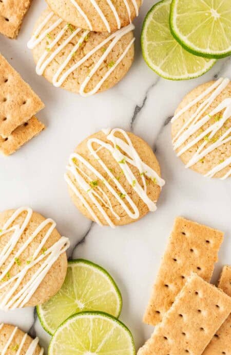 Overhead shot of key lime pie cookies drizzled with white chocolate glaze, surrounded by lime slices and graham crackers