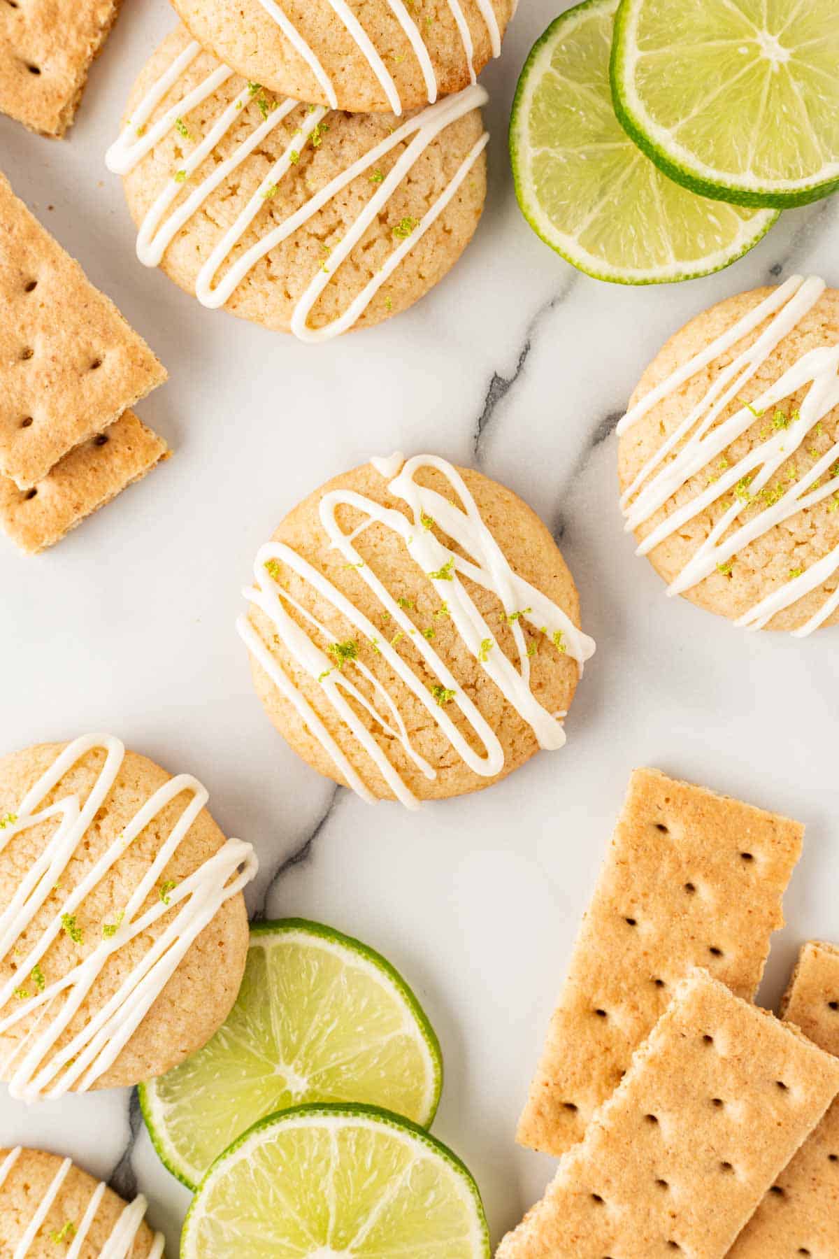 Overhead shot of key lime pie cookies drizzled with white chocolate glaze, surrounded by lime slices and graham crackers