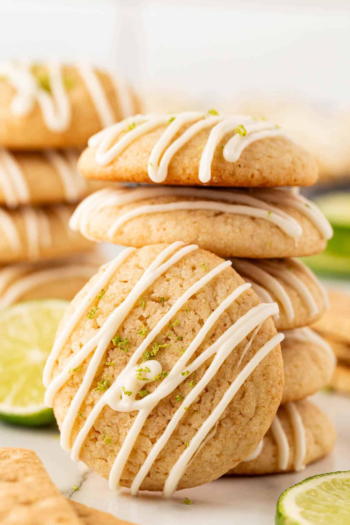 Stack of iced key lime pie cookies with lime zest on top and more cookies blurred in the background