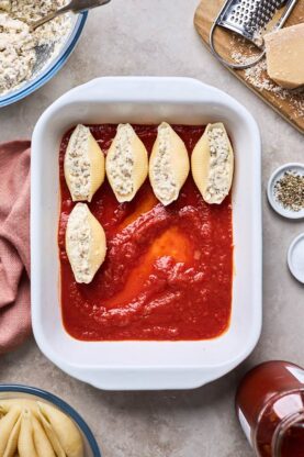 A white casserole dish with tomato sauce on the bottom and filled shells lining the pan