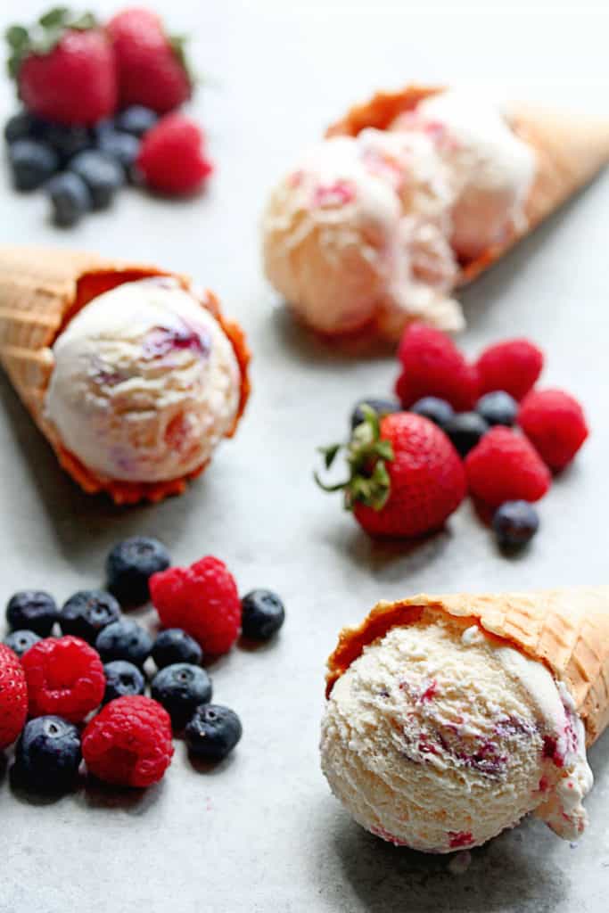 Three waffle cones containing one scoop of berry ripple ice cream with fresh berries all around