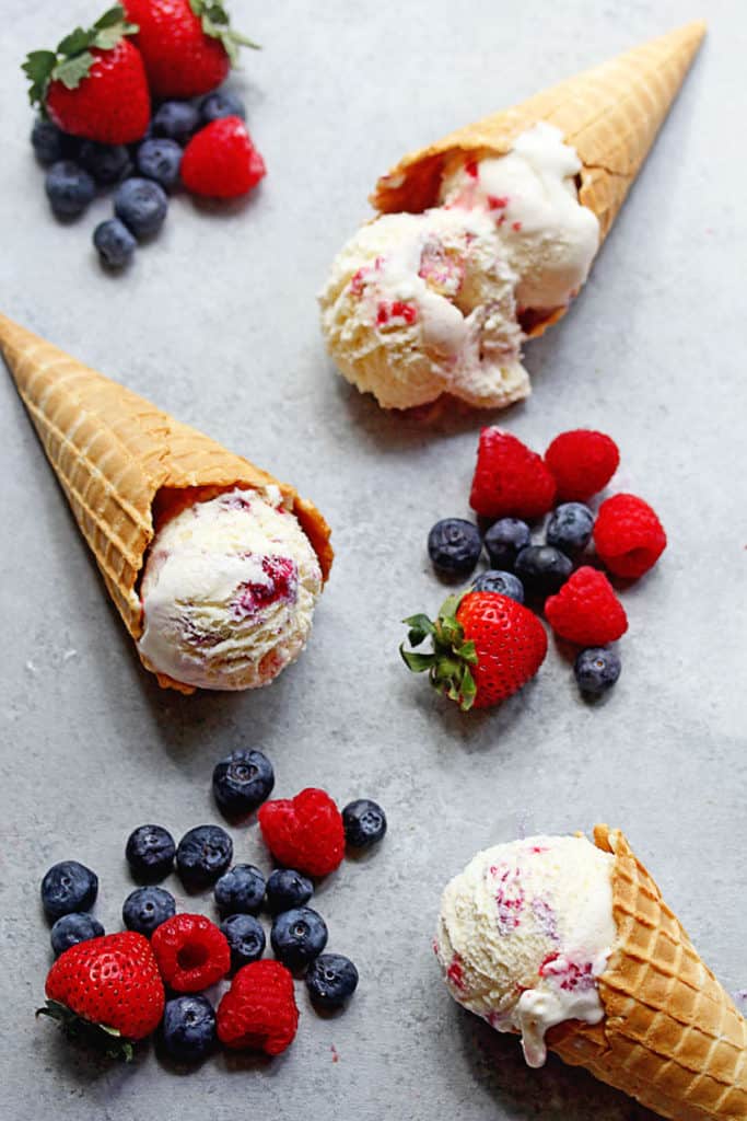 Overhead shot of three waffle cones containing one scoop of berry ripple ice cream with fresh berries all around