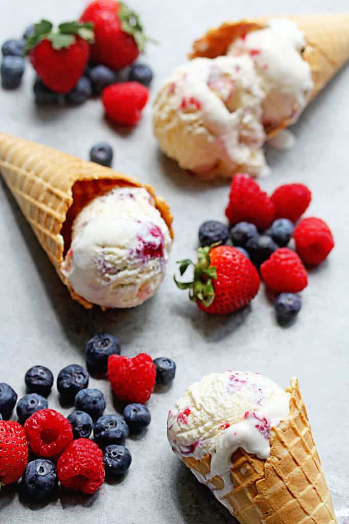 Three waffle cones containing one scoop of melting berry ripple ice cream with fresh berries all around