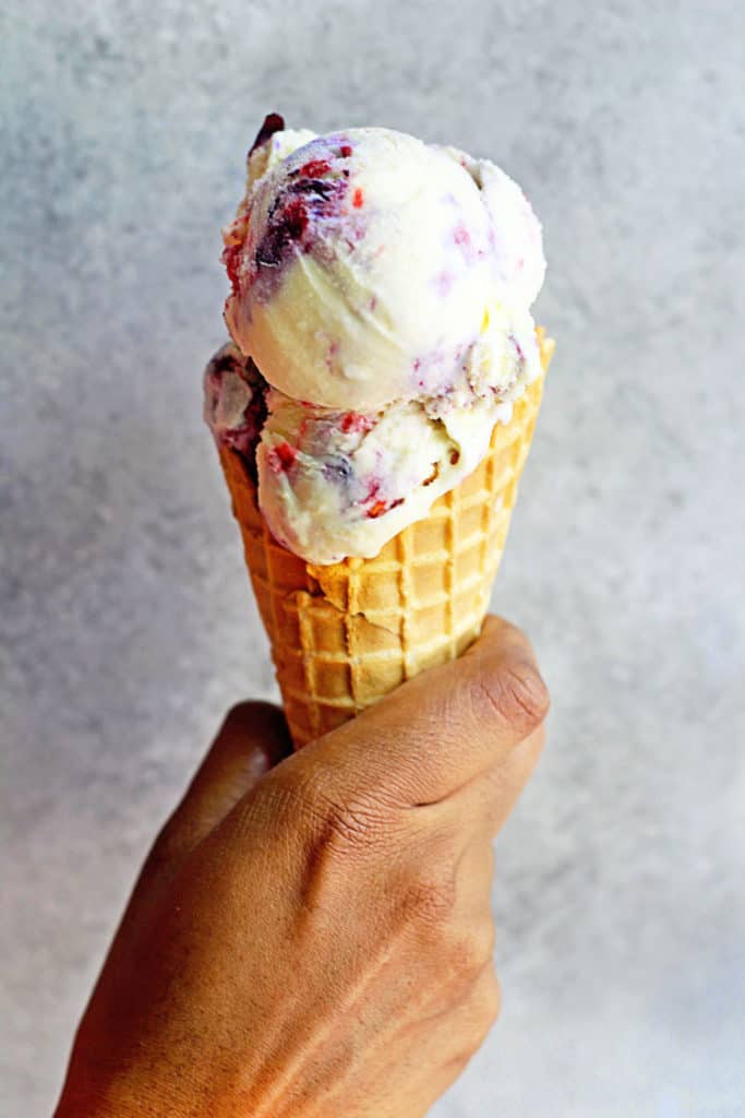 A hand holding a waffle cone containing scoops of creamy berry ripple ice cream 