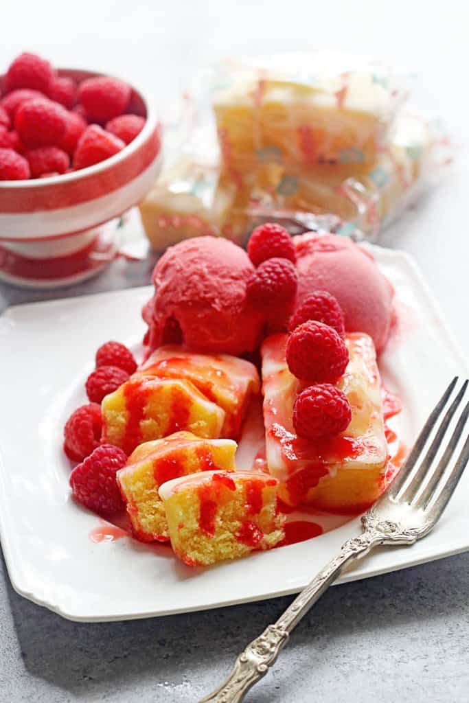 Lemon Raspberry Poke Cake with a fork sitting on a square, white plate with raspberry sorbet and topped with fresh raspberries and a bowl of raspberries and poke cake packages in the background