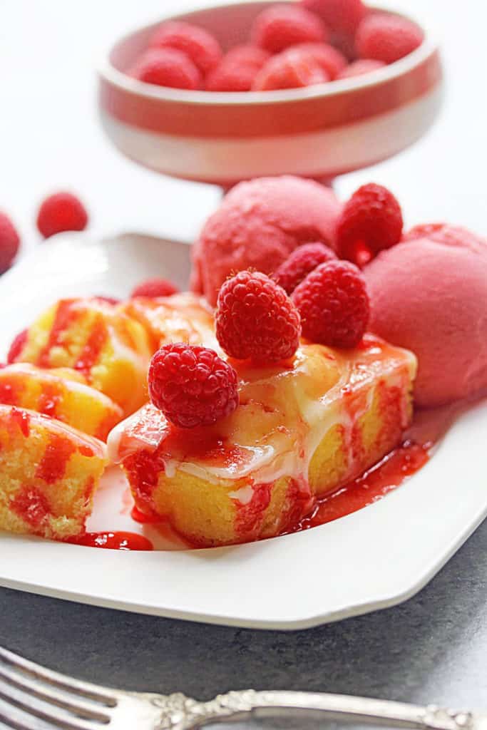 Close up of Lemon Raspberry Poke Cake sitting on a square, white plate with raspberry sorbet and topped with fresh raspberries and a bowl of raspberries in the background