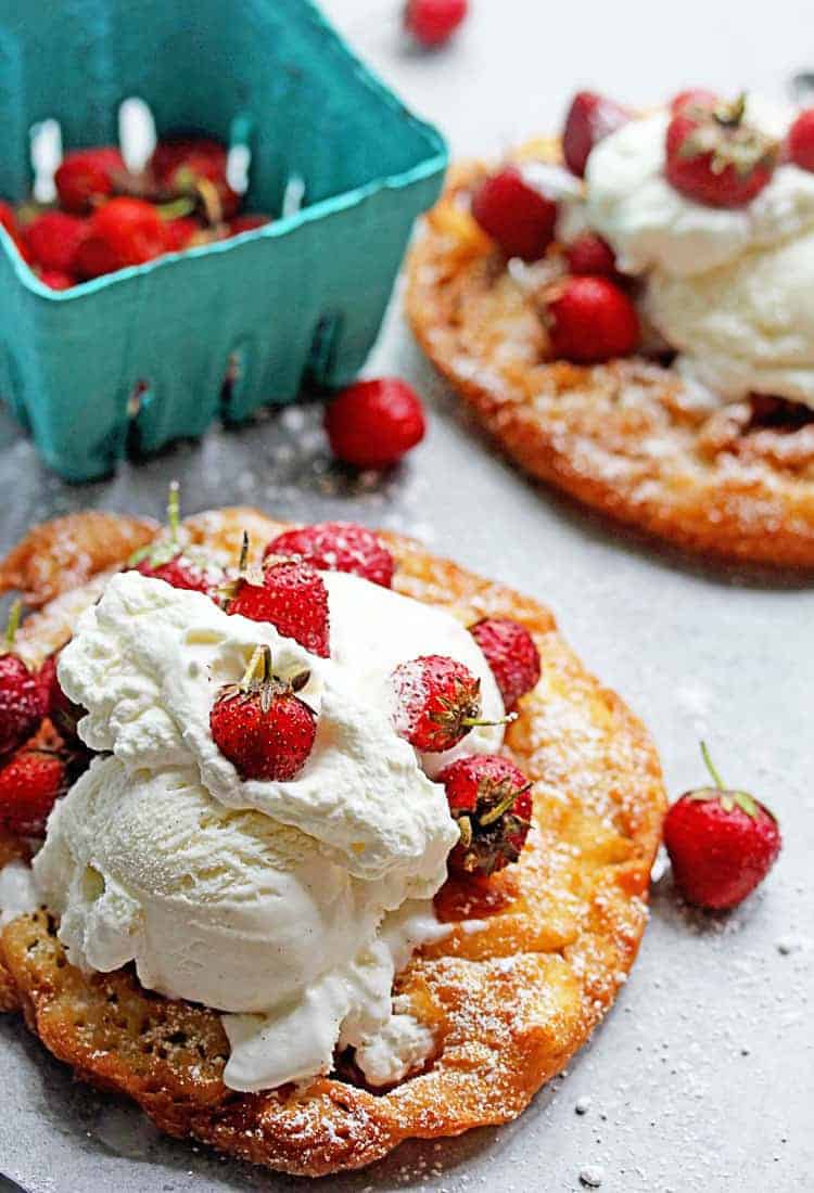 Close up of strawberry shortcake funnel cake topped with powdered sugar, whipped cream and fresh strawberries with a basket of strawberries and another funnel cake in the background. 