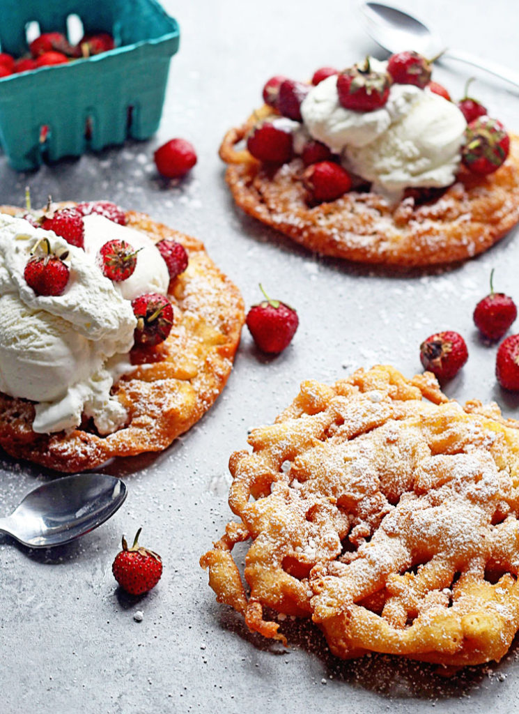 Strawberry Shortcake Funnel Cake Recipe with three funnel cakes displayed topped with powdered sugar, whipped cream and strawberries.
