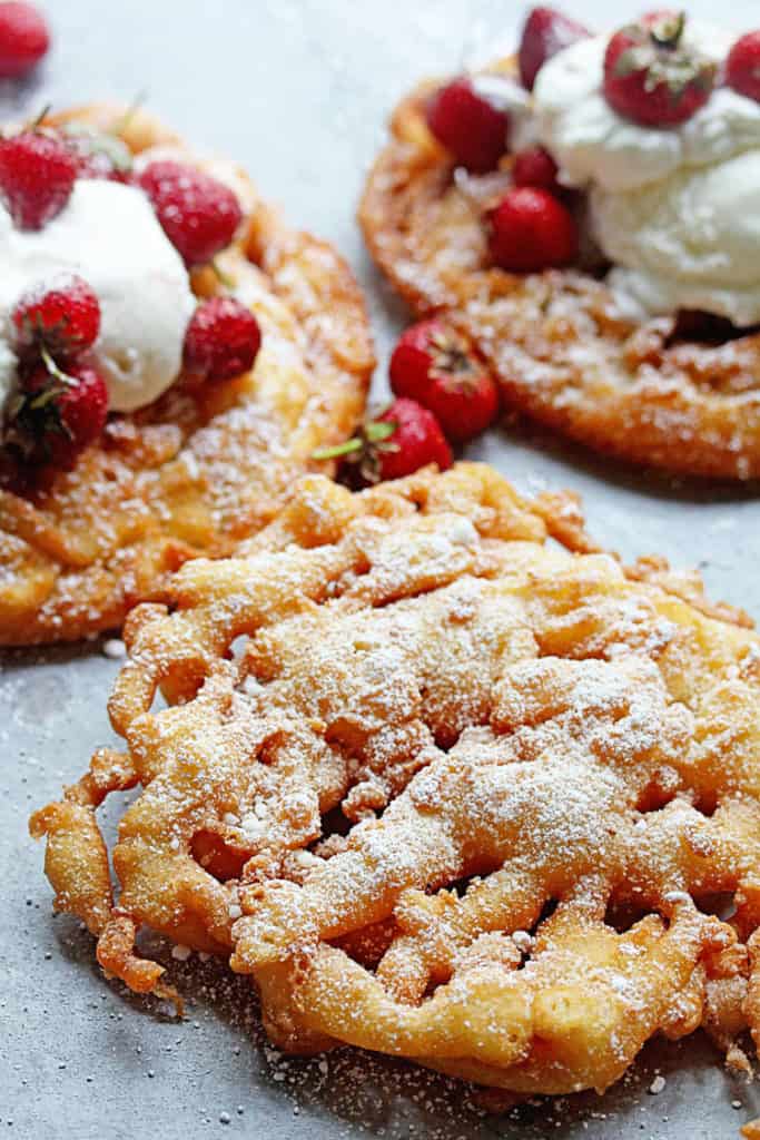Funnel Cake Recipe displayed with three funnel cakes. Two in the background are topped with whipped cream and strawberries, and the one in the foreground is topped with powdered sugar.