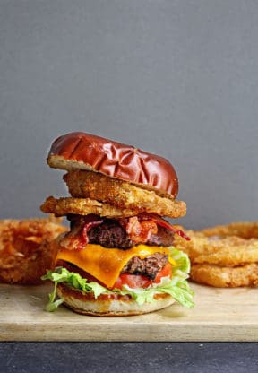 Ultimate Bacon Cheddar Burger served with crispy fried onion rings on a wooden cutting board