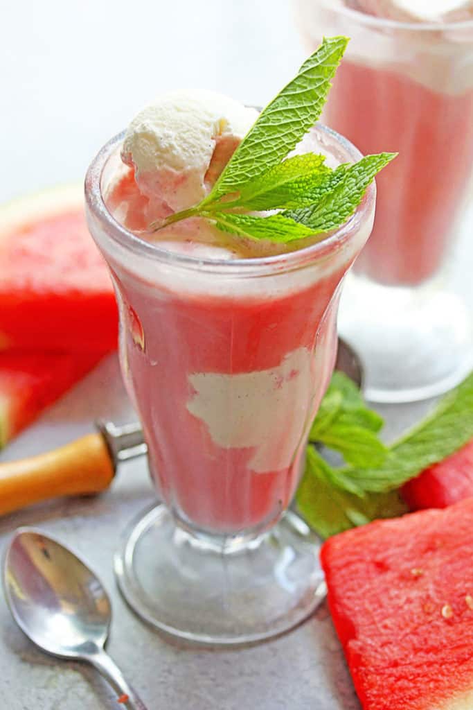 Close up of one watermelon ice cream float contained in a clear glass and topped mint with spoons and watermelon wedges near by and other float in the background