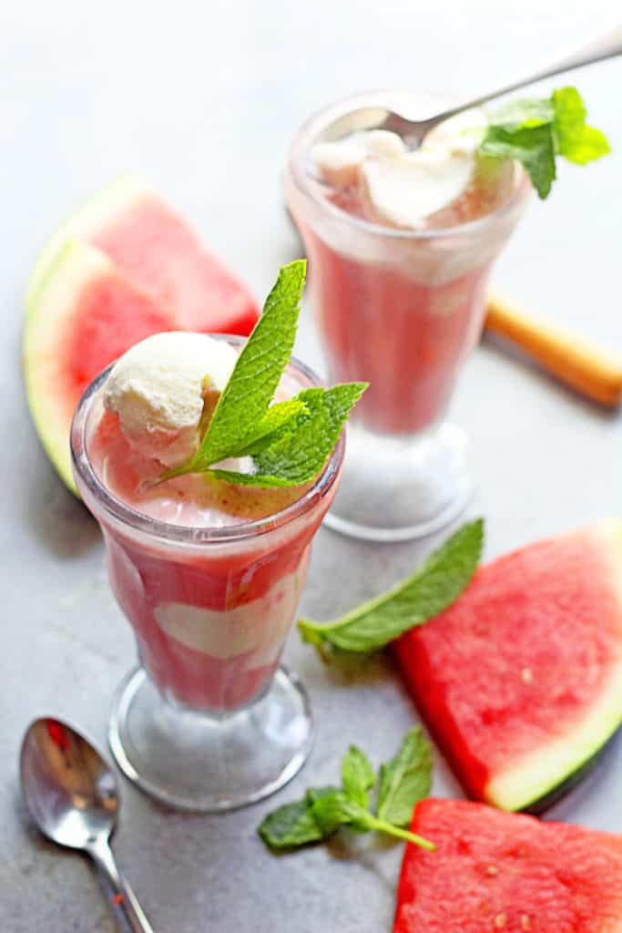 Two watermelon ice cream floats contained in clear glasses and topped mint with spoons and watermelon wedges near by