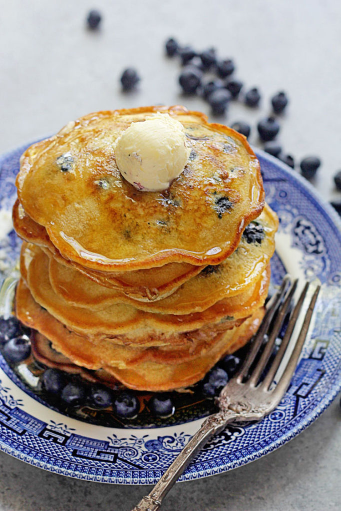 Easy blueberry pancakes stacked high on a blue plate with butter and fresh blueberries surrounding the plate