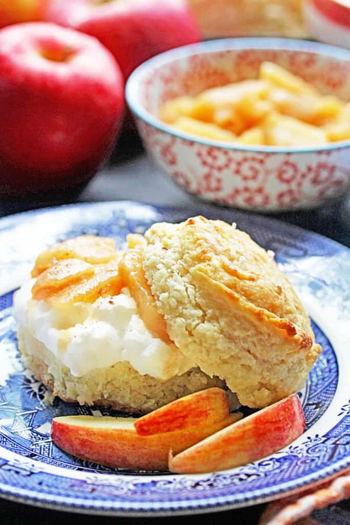 A Brown Sugar Biscuit Apple Shortcake on a blue and white plate with apple slices and whole apples and a bowl of apple pieces in the background