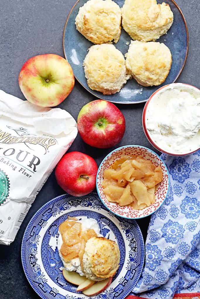 Overhead shot of a Brown Sugar Biscuit Apple Shortcake on a blue and white plate with apple slices and whole apples, a bag of flour, biscuits and a bowl of apple pieces next to it