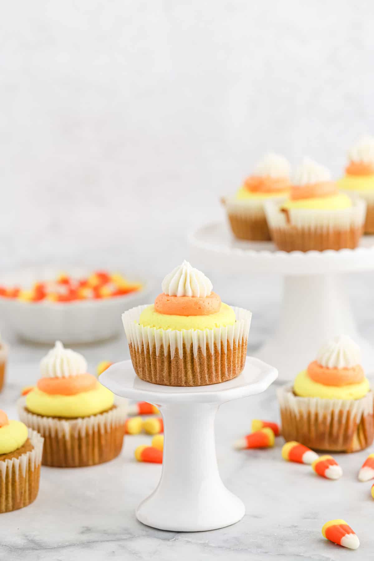 Pumpkin cupcakes with candy corn buttercream on a white cake stand with more in background.