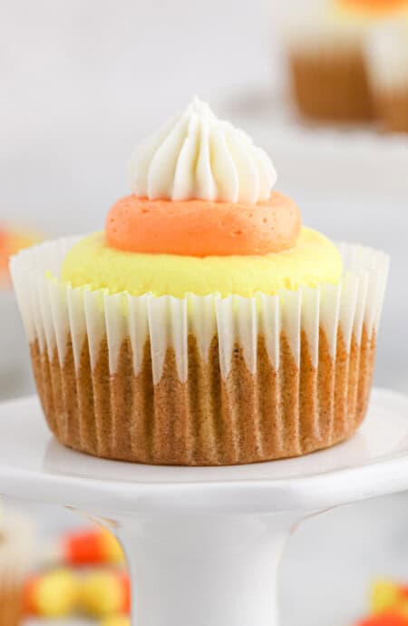 Pumpkin cupcake with candy corn frosting on a cake stand with more in back.
