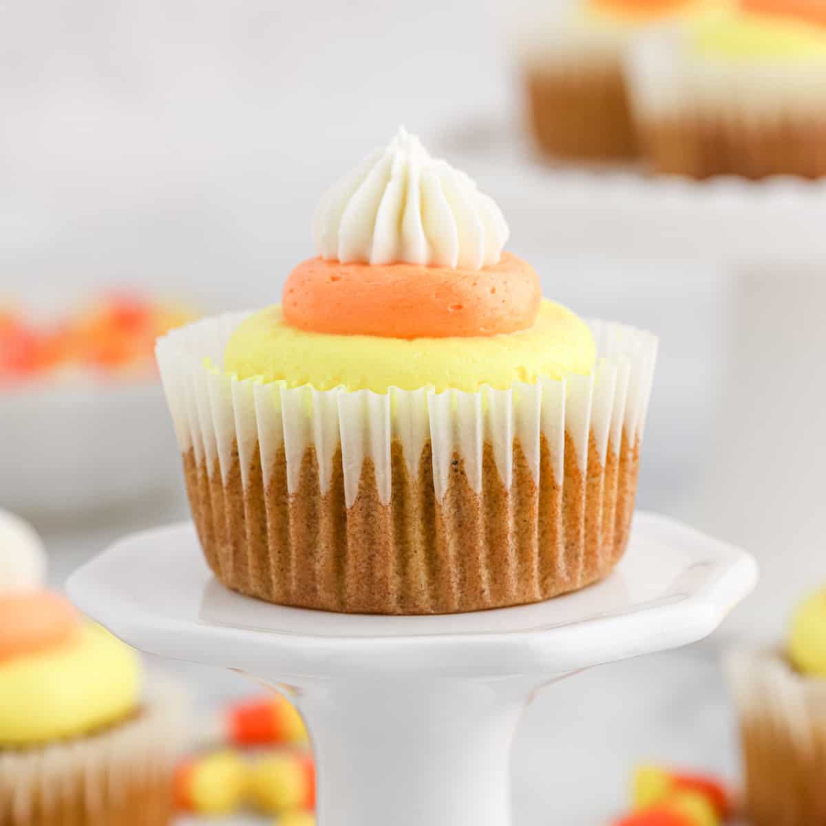 A Pumpkin Cupcake with Candy Corn Buttercream with candy corn pieces next to it and more cupcakes and a pumpkin in the background 