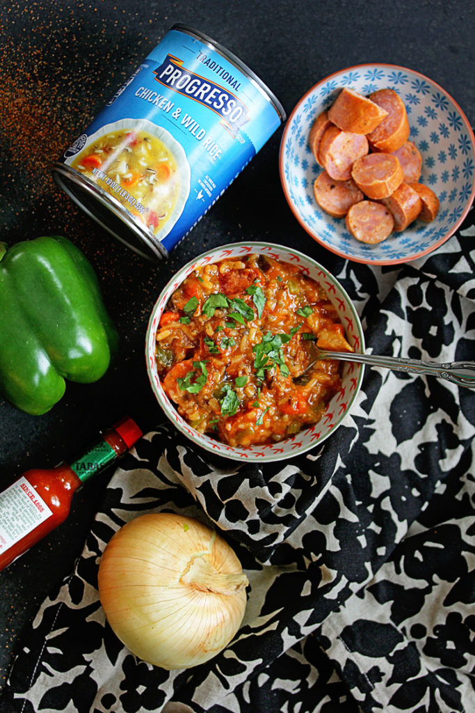 Overhead shot of jambalaya soup with a can of Progresso soup, sliced sausage, a bell pepper, hot sauce and an onion by it