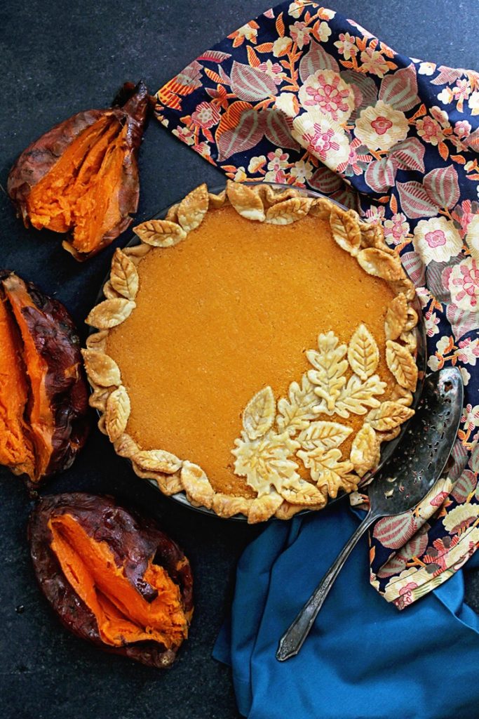 An overhead of a beautifully decorated sweet potato chess pie with roasted sweet potatoes and napkins surrounding it