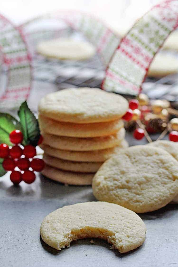Close up of a stack of Butter Ricotta Cookies along with one with a bite taken out of it with more cookies on a cooking rack with a Christmas ribbon in the background