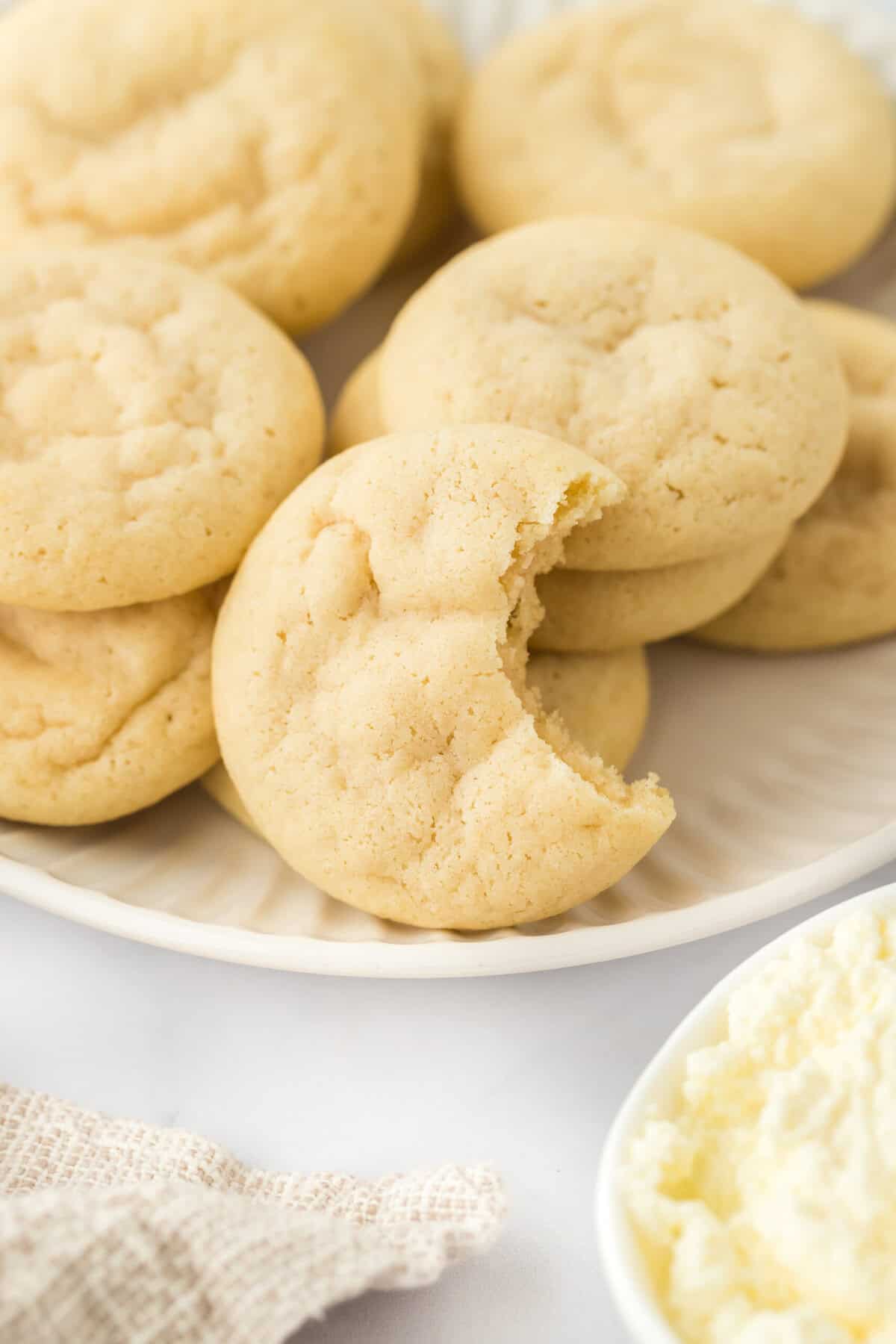 A ricotta cookie recipe on a white plate with ricotta in a bowl with a bite inside of it.