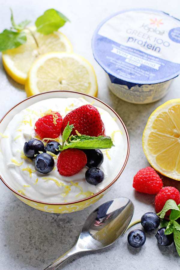 A bowl full of easy lemon mousse topped with fresh berries and mint with lemon slices, raspberries, blueberries, a package of yogurt and a spoon next to it
