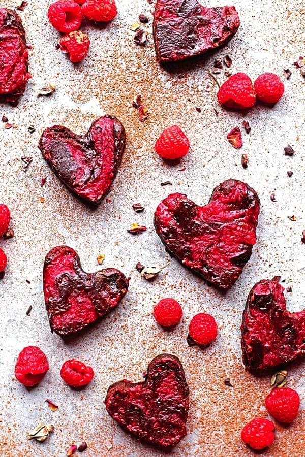 Heart shaped raspberry brownies on a plain background with raspberries surrounding them