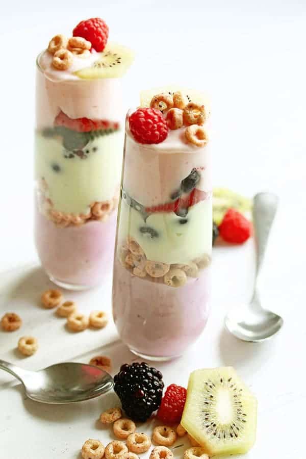 Two cereal parfaits with silver spoons, berries and Cheerio cereal pieces lying next to them.