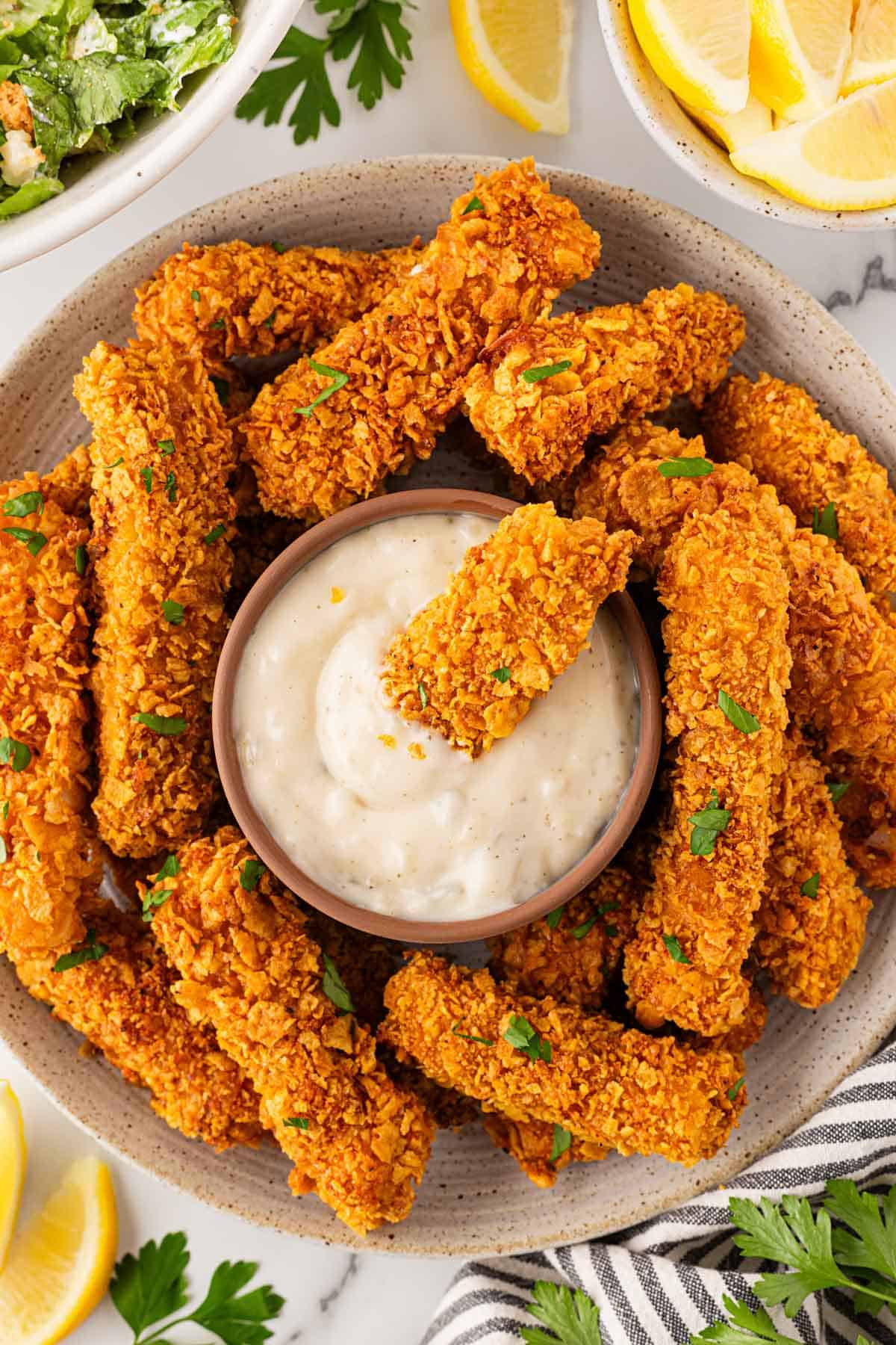 Overhead shot of a golden baked fish stick dipped into creamy tartar sauce surrounded by more baked fish sticks on the plate