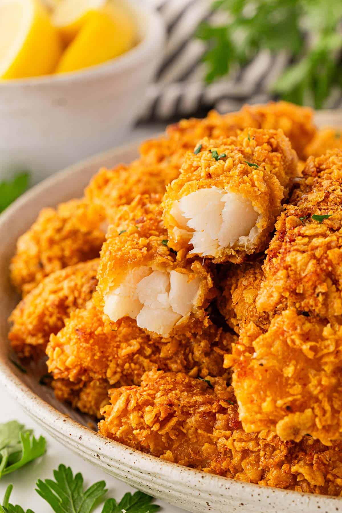 Closeup of baked fish sticks stacked in a bowl, showing the tender flaky white fish interior, with lemon wedges and parsley in the background