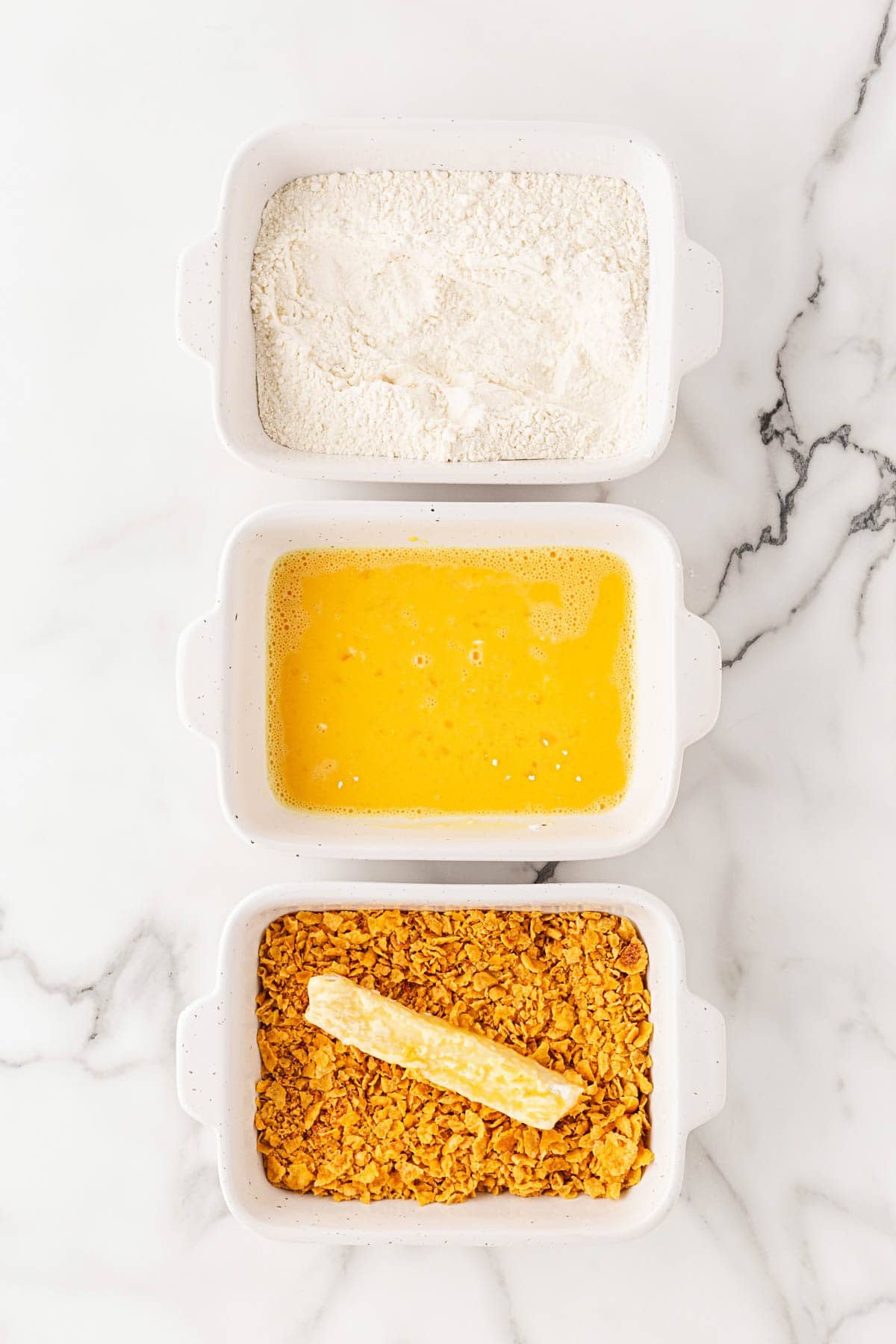 Dredging station setup with flour, beaten eggs, and crushed cornflakes arranged in three bowls, with a fish strip being coated in the cornflake mixture