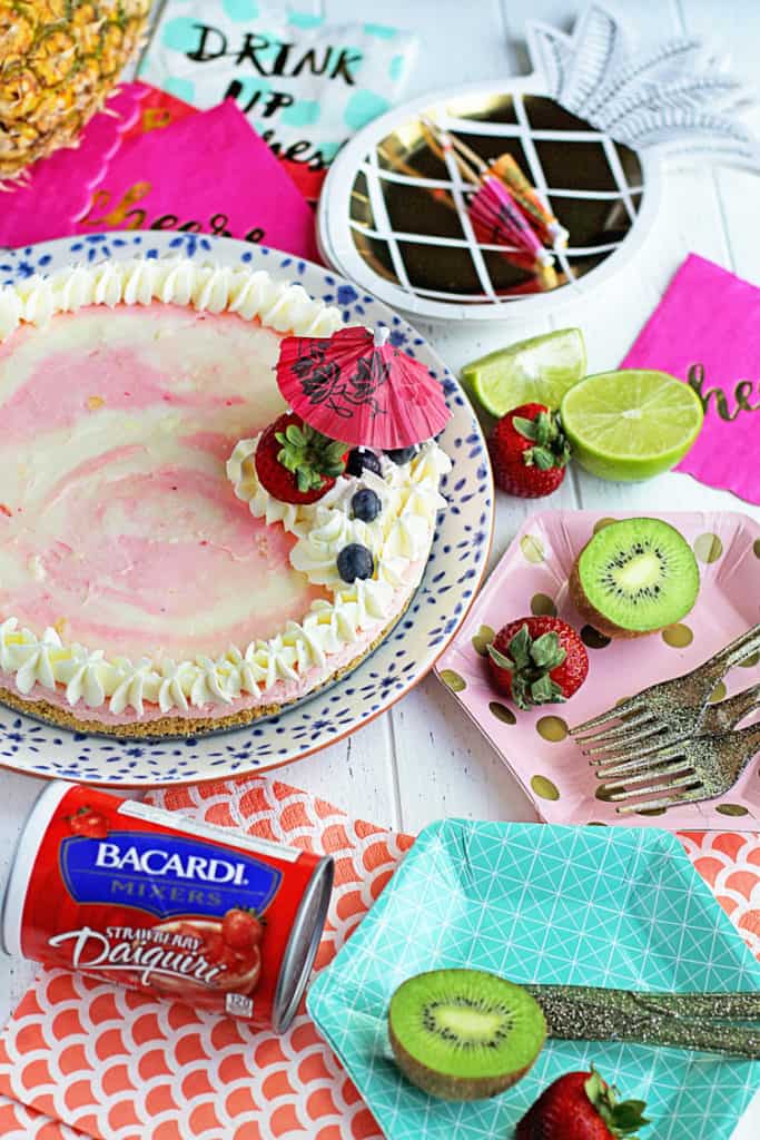 Bacardi Miami Vice No Bake Cheesecake with colorful napkins, plates, utensils, a bottle of frozen bacardi daquiri mix, a whole pineapple and other pieces of fruit