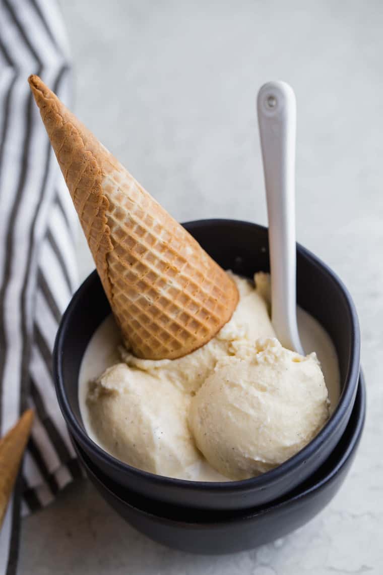 A close up of vanilla bean ice cream in a large black bowl with waffle cone and spoon ready to serve