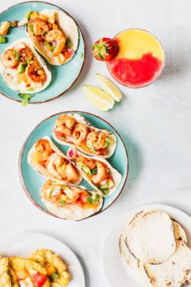 Plates of jerk shrimp tacos on blue plates with limes, tortillas and drinks ready to serve