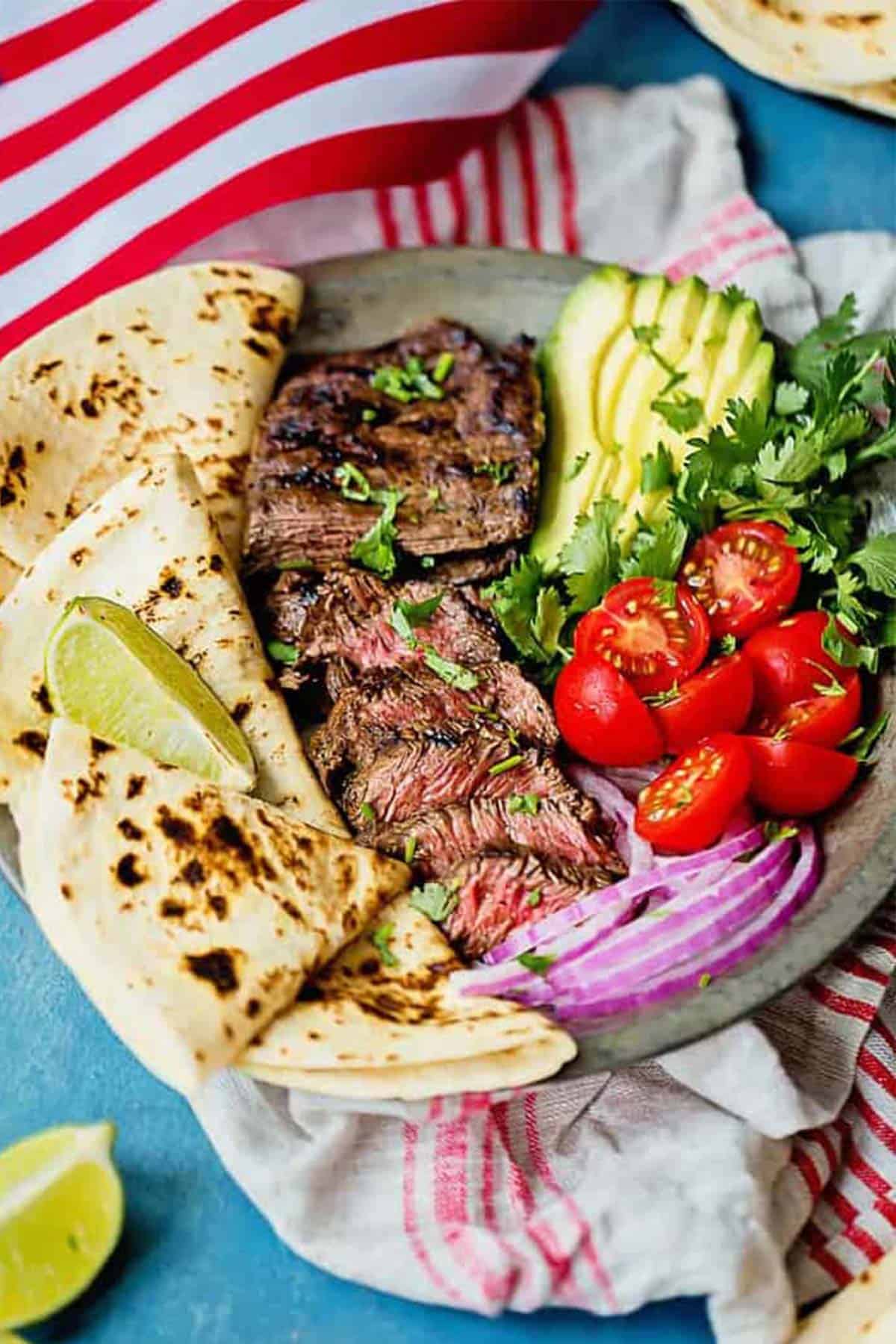 Carne asada recipe sliced on a platter with tortillas and limes scattered around