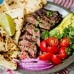 Grilled carne asada steak sliced on a plate with tomatoes, avocado and flour tortillas.