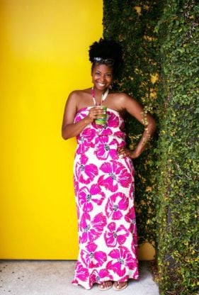 Jocelyn standing in a purple flower printed sleeveless dress holding a drink with a straw in front of a yellow wall