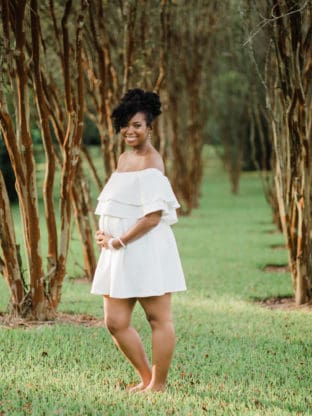 A pregnant Jocelyn Delk Adams holds her belly dressed in a white maternity dress