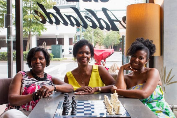 Jocelyn with her mom, center, and aunt in New Orleans seated behind a chess board and pieces